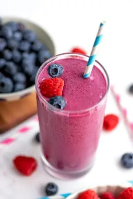 A raspberry blueberry smoothie on a white table with raspberries and blueberries separated into two bowls next to the smoothie.
