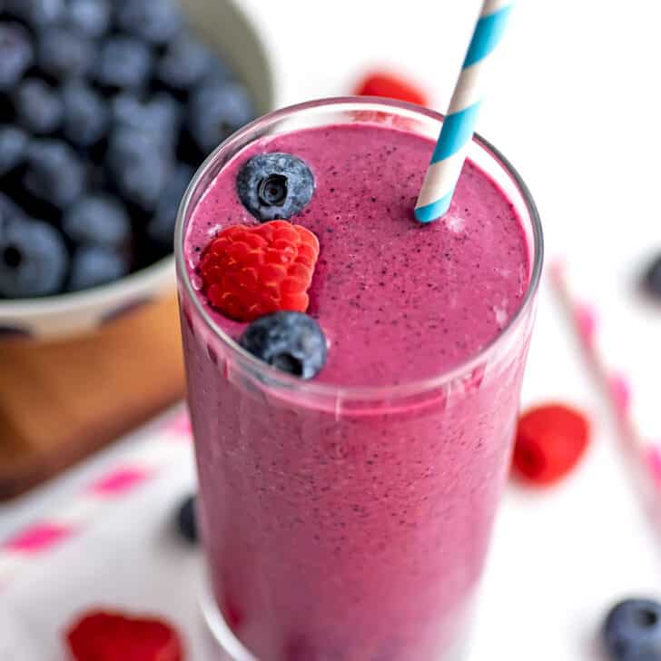 A raspberry blueberry smoothie on a white table with raspberries and blueberries separated into two bowls next to the smoothie.