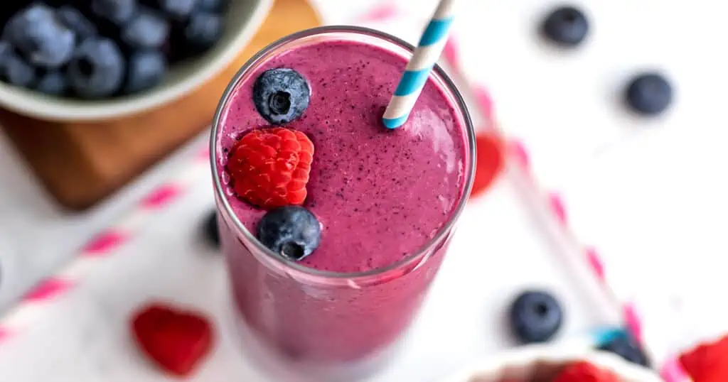 A raspberry blueberry smoothie with a white and blue paper straw and blueberries and raspberries on top.
