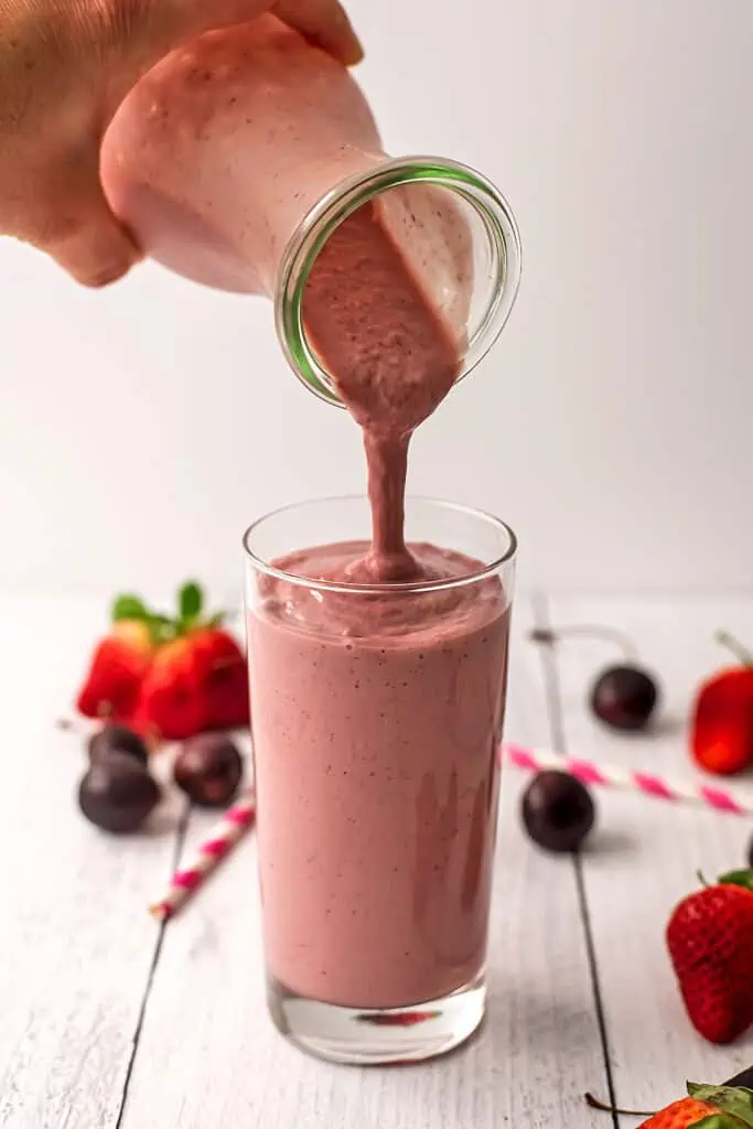 A strawberry cherry smoothie being poured into a glass. Cherries and strawberries are out of focus in the background.