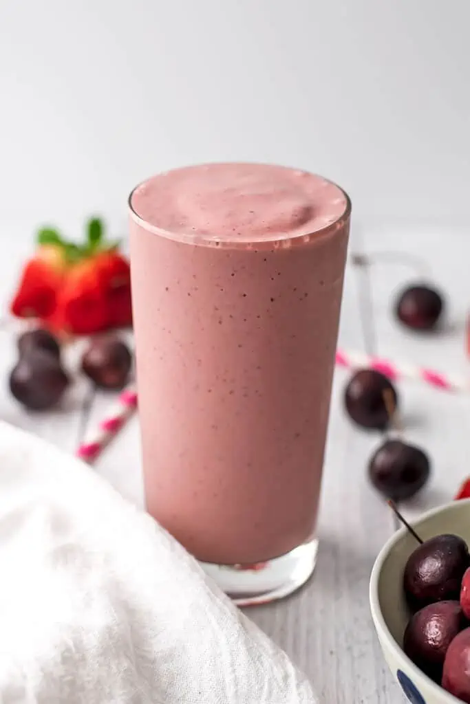 A cherry strawberry smoothie in a tall glass with cherries and strawberries out of focus behind the smoothie.