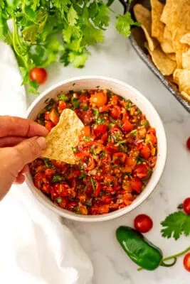Cherry tomato salad in a white bowl with a chip being dipped into the salsa.
