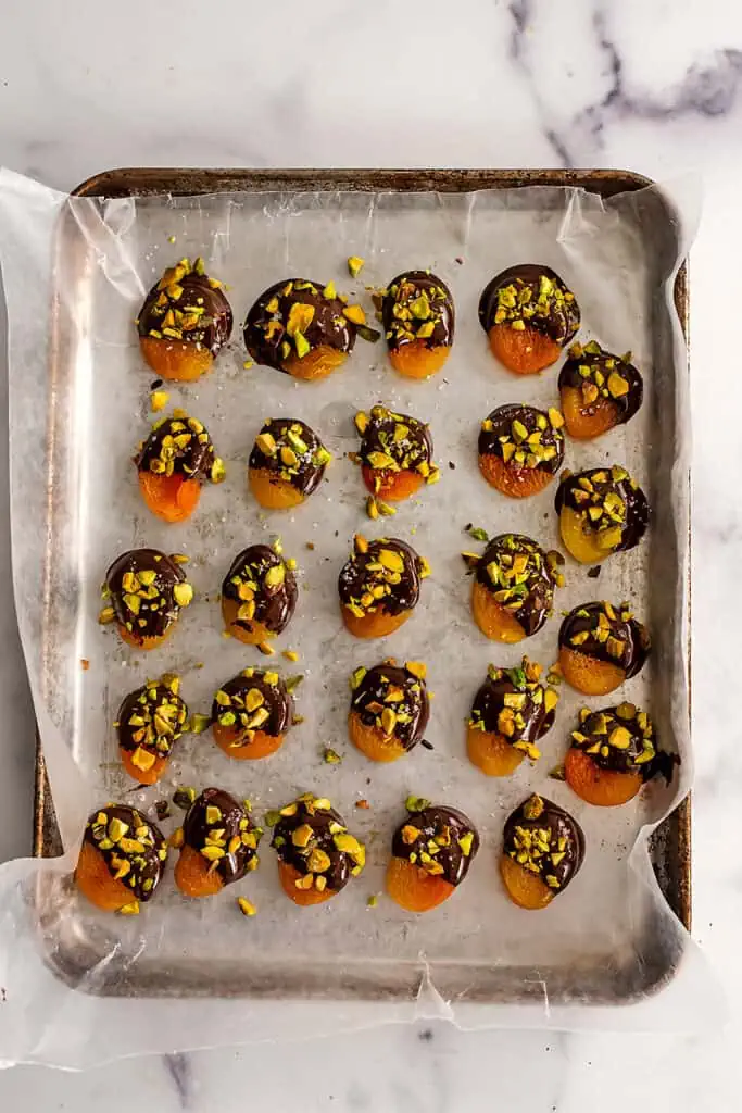 Dried apricots dipped in chocolate and sprinkled with pistachios on a baking sheet.