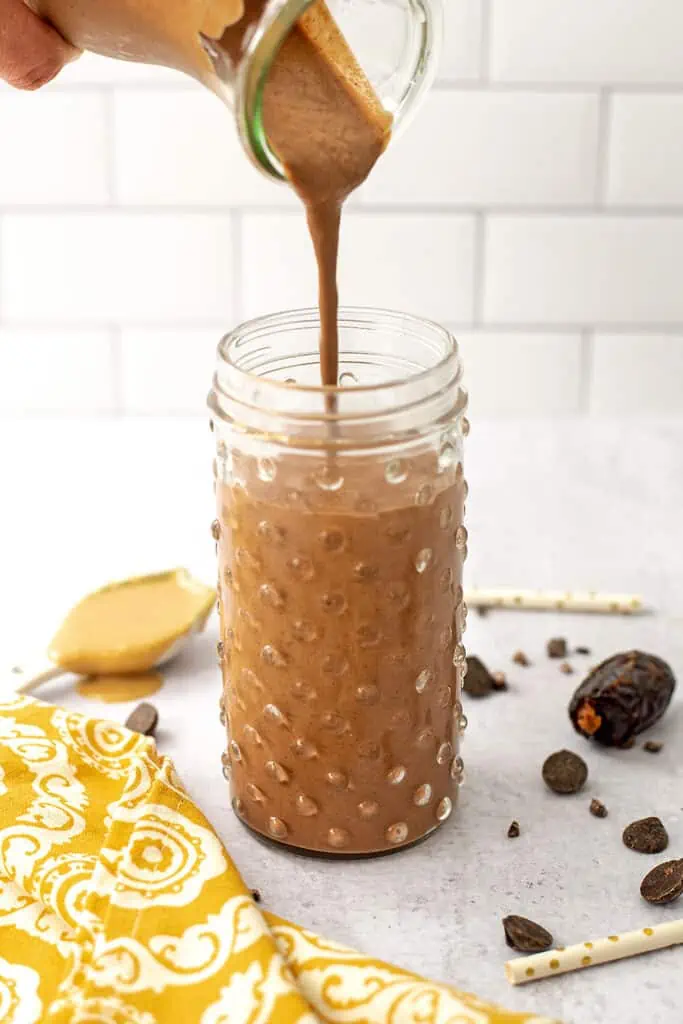 Chocolate Tahini Smoothie being poured into a tall glass. Medjool dates and chocolate chips are scattered on the table. 