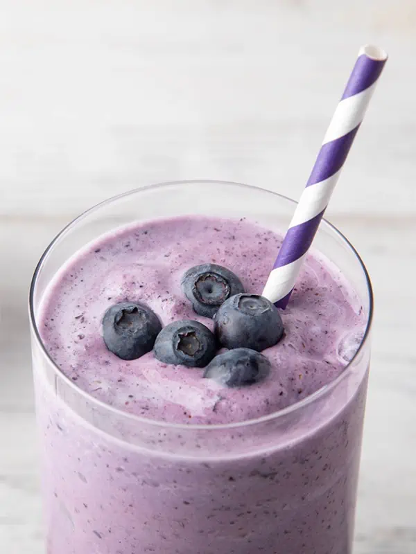 A blueberry protein smoothie in a glass with blueberries on top with a purple and white straw.
