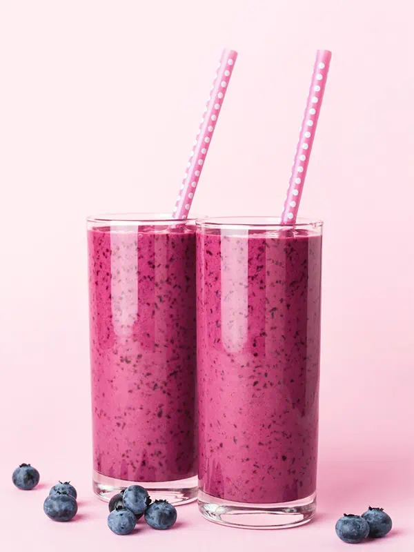 Two protein smoothies in glasses side by side with matching straws. Blueberries are laying across the table around the smoothies.