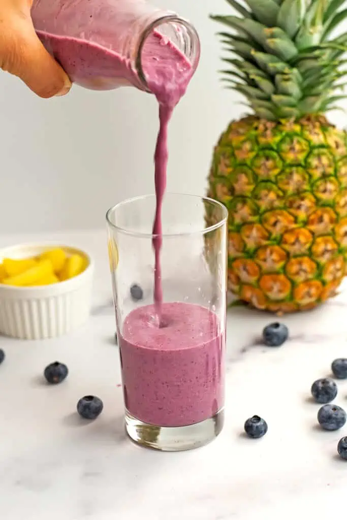 A pineapple blueberry smoothie being poured into a glass. Blueberries and a whole pineapple are sitting on the table next to the smoothie.