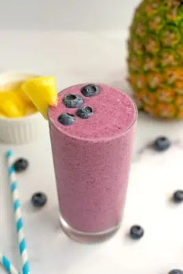 A pineapple blueberry smoothie in a tall glass sitting on a white table with blueberries.