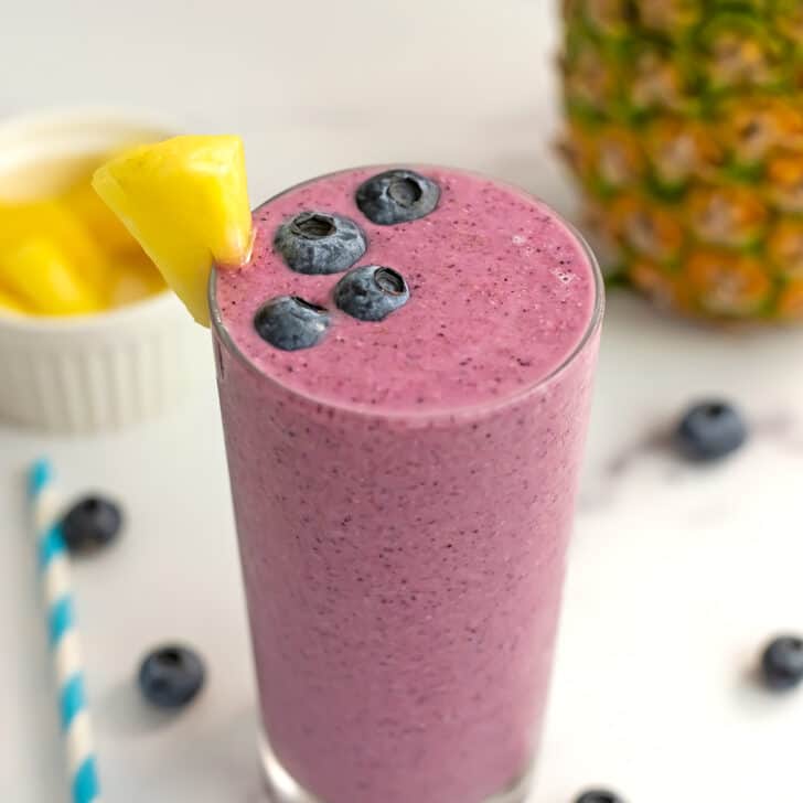 A pineapple blueberry smoothie in a tall glass sitting on a white table with blueberries.