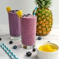 Pineapple blueberry smoothies on a white table with blueberries and pineapples on top.