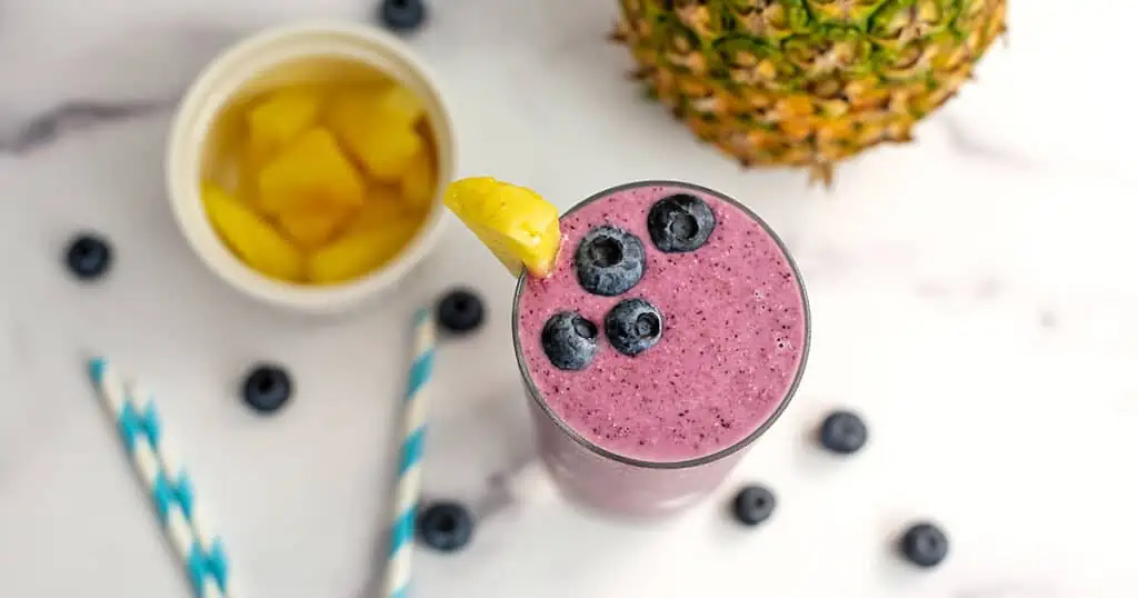 An overhead view of a pineapple blueberry smoothie. Pineapple chunks are in a white ramekin sitting next to the smoothie and blueberries are scattered around the table.