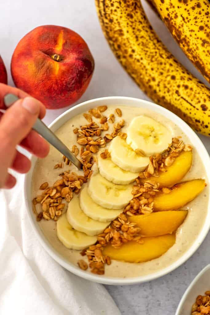 Banana peach smoothie bowl recipe with granola, banana and peaches. A hand is reaching for the spoon.