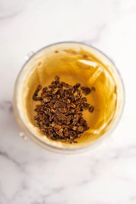 Cookie dough smoothie bowl in a blender cup with cacao nibs being added.