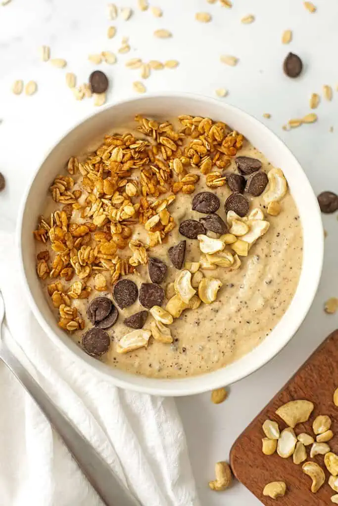 Cookie dough smoothie bowl on a white table with granola and chocolate chips spread around it.