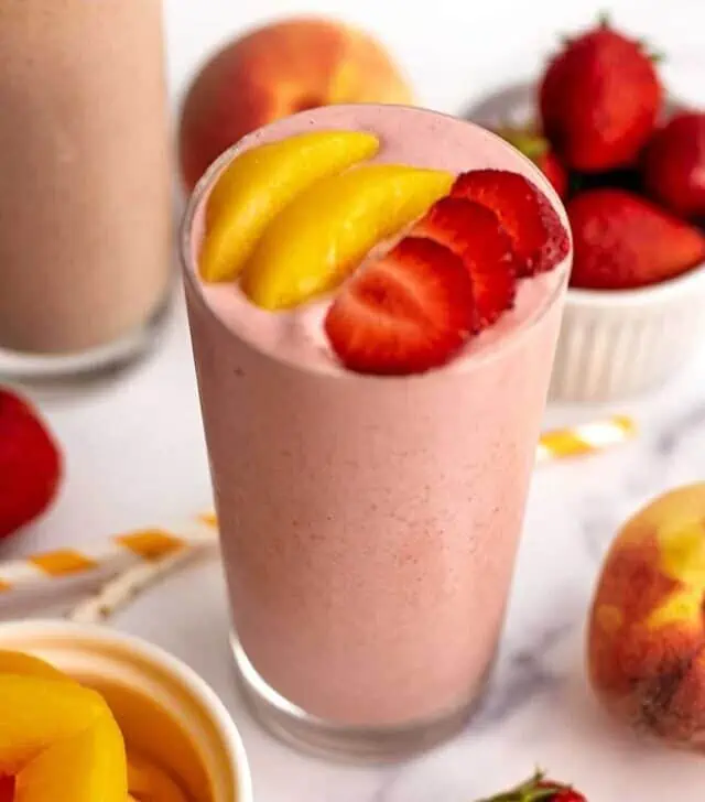Strawberry peach smoothie with sliced peaches and strawberries on top surrounded by strawberries on a white table.