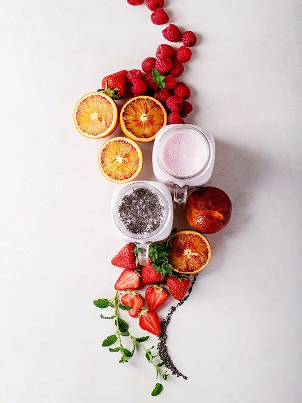 Two smoothies surrounded by fruits such as blueberries, strawberries, raspberries and blood oranges.