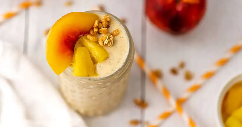 Peach and pineapple smoothie with peaches and pineapple chunks and granola on top. Straws are sitting on a white table next to it.
