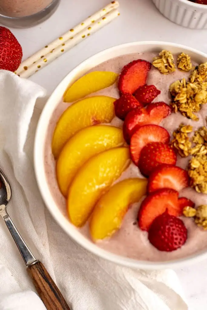 Peach and strawberry smoothie in a bowl lined with sliced peaches, strawberries and bunches of granola on a white tablecloth. 