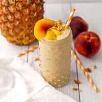 Pineapple peach smoothie with peaches and a straw sitting on a white table.