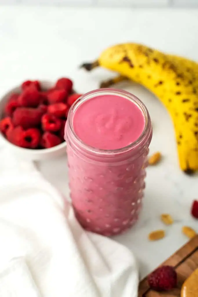 Peanut butter raspberry smoothie in a glass with raspberries and banana behind it.