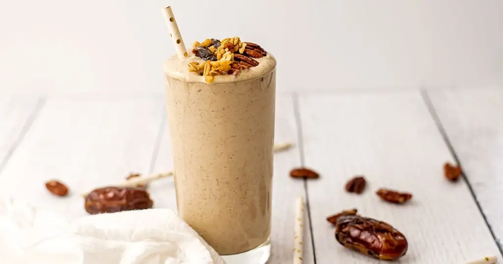 Pecan pie smoothie with pecans, dates, and granola on top with a straw on a white table.