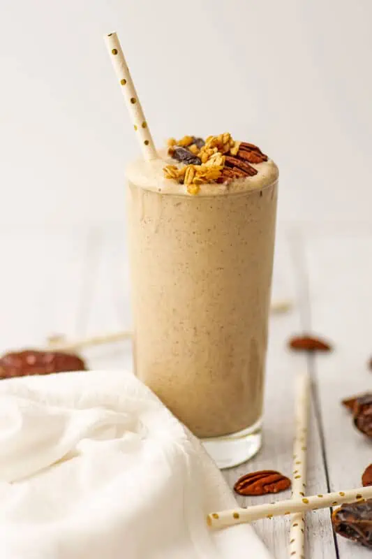 Pecan smoothie with pecans, dates, and granola on top with a straw on a white table.
