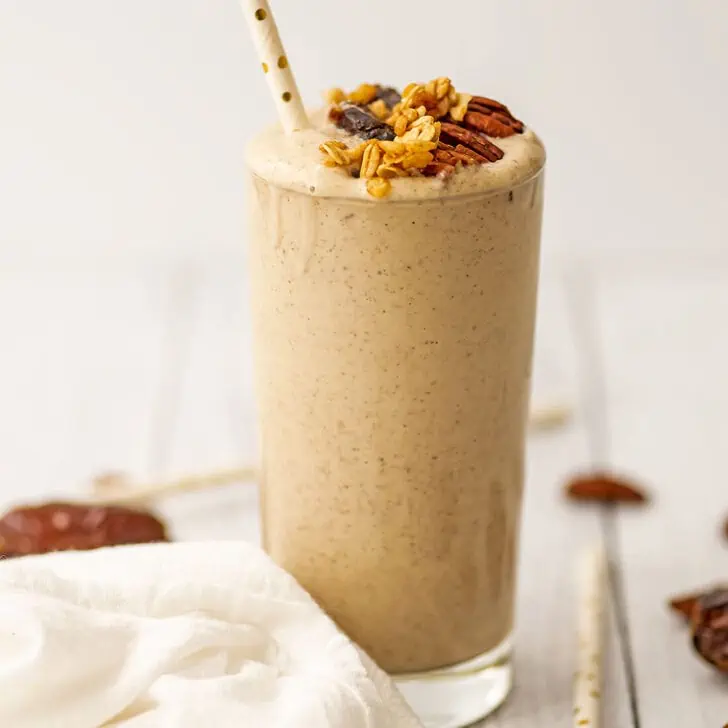Pecan smoothie with pecans, dates, and granola on top with a straw on a white table.