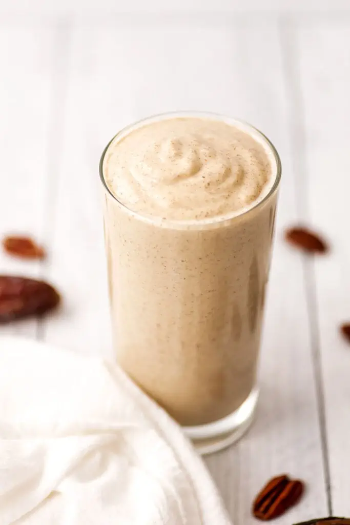 Pecan smoothie in a large glass on a white table with pecan pieces laying around the smoothie.
