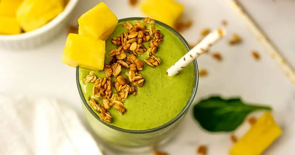 Pineapple matcha smoothie with granola and pineapple wedges on the side sitting on a white table.