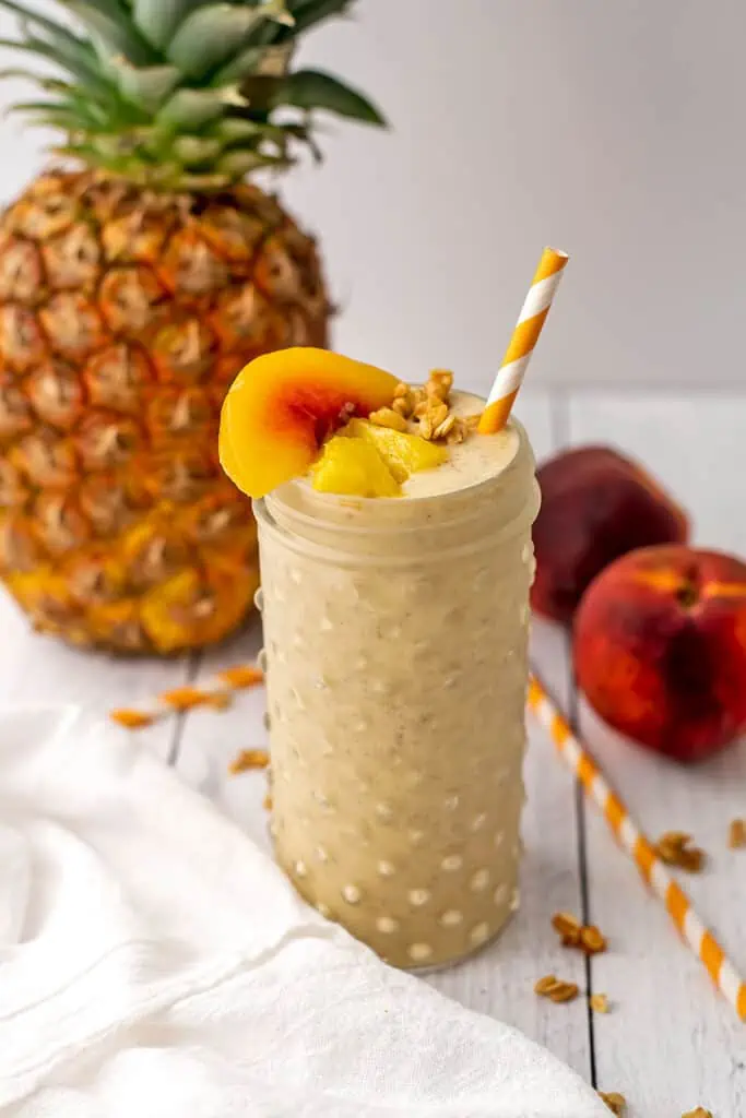 Pineapple peach smoothie in a mason jar with peaches and pineapple on top with a straw. A pineapple is out of focus in the background.