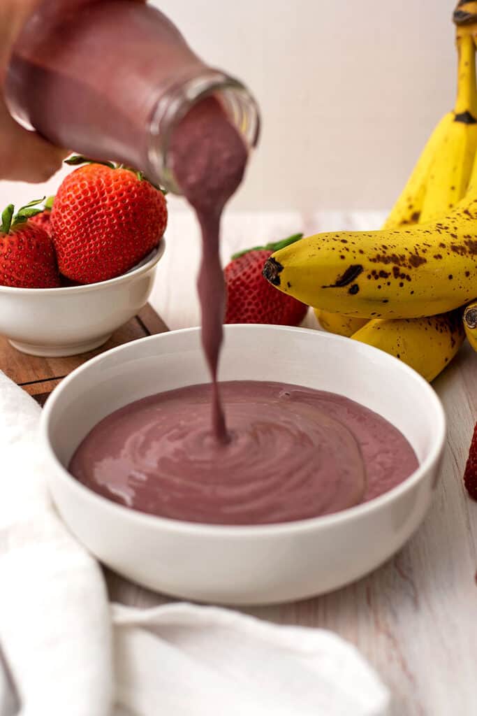 Strawberry and banana acai bowl being poured into a white bowl.
