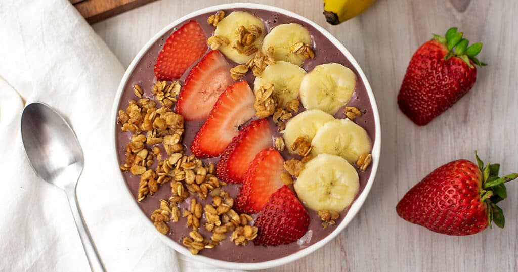 Strawberry banana acai bowl with a spoon. There are sliced strawberries, bananas, and granola on top of the acai smoothie bowl.