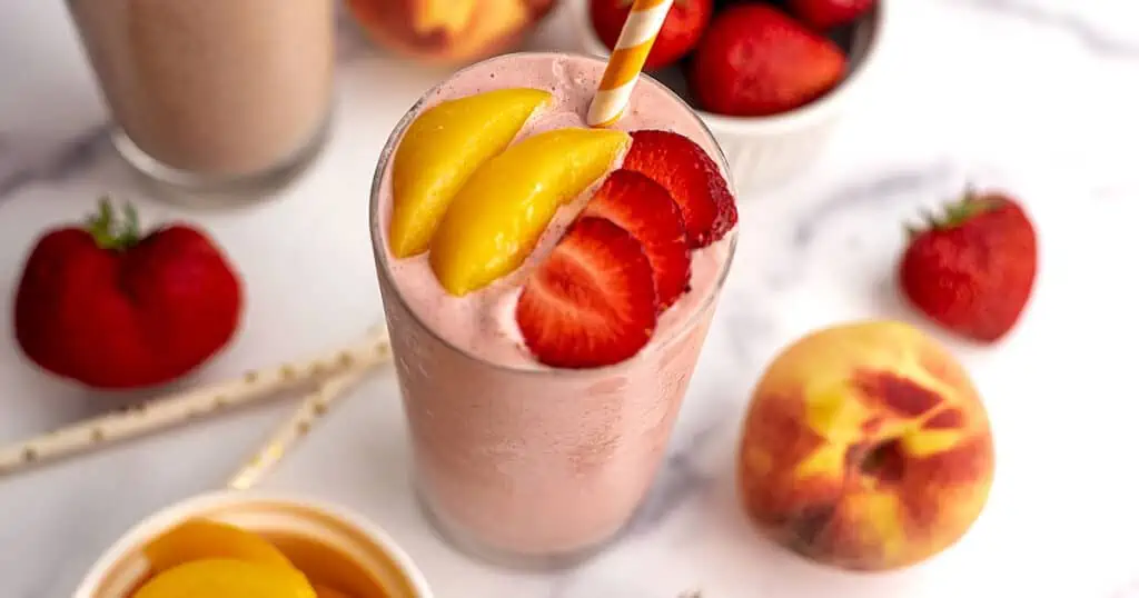 Strawberry peach smoothie no yogurt on a white table surrounded by peaches and strawberries.