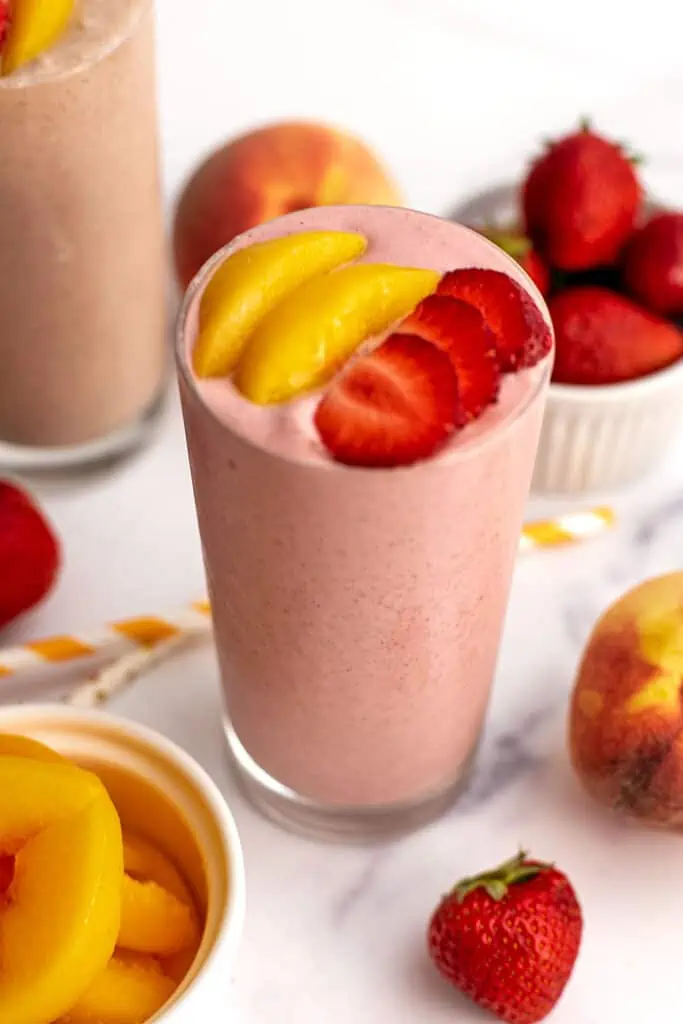 Strawberry peach smoothie with sliced peaches and strawberries on top surrounded by strawberries on a white table.