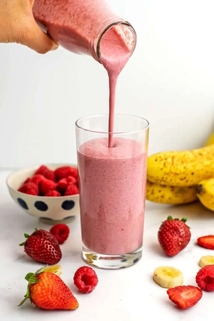 Strawberry raspberry and banana smoothie being poured into a glass with strawberries, bananas and raspberries in the background.