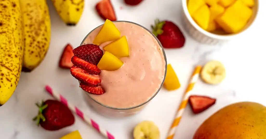 Mango strawberry banana smoothie on a white table with mangos, bananas, and strawberries laying around the glass.