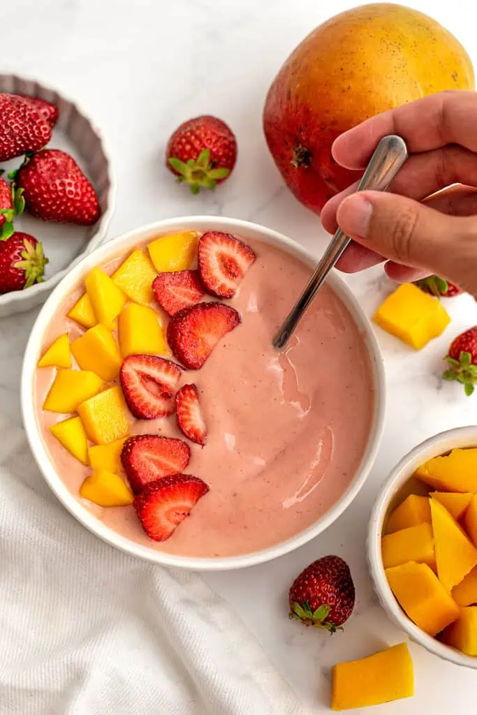 Mango strawberry smoothie bowl in a bowl with fresh fruit. A hand is grabbing the spoon.