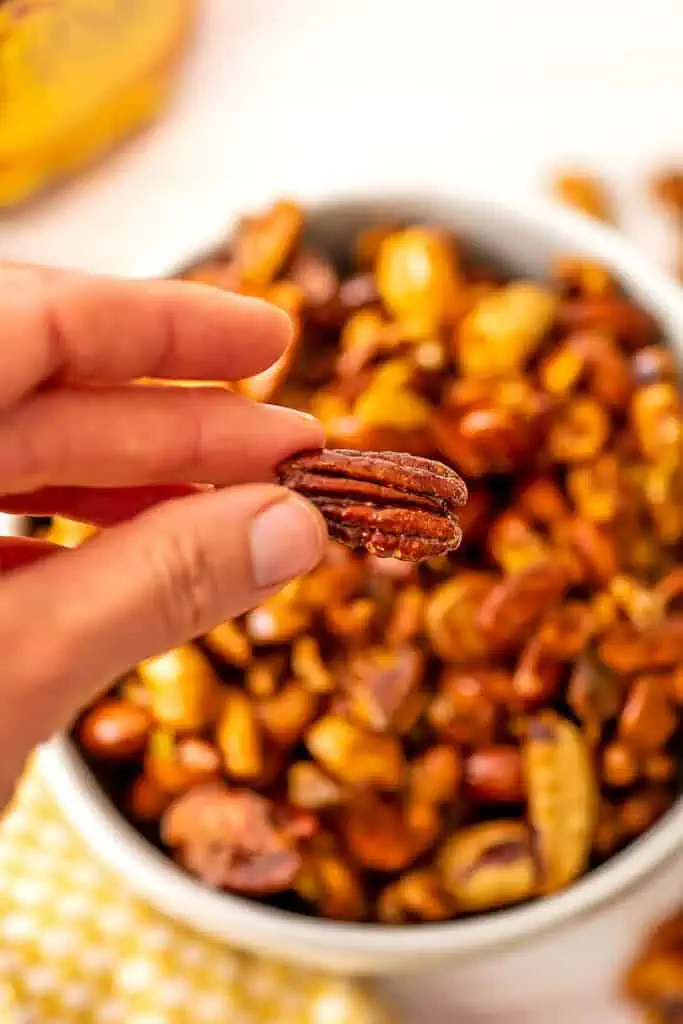 Hand holding a maple roasted pecan over a bowl of nuts.