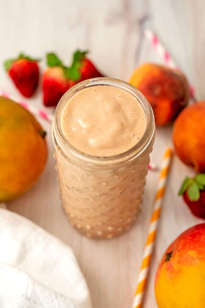 Peach mango strawberry smoothie in a tall glass with strawberries and peaches in the background.