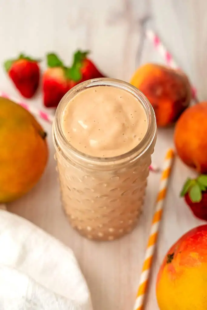 Peach mango strawberry smoothie in a tall glass with strawberries and peaches in the background.