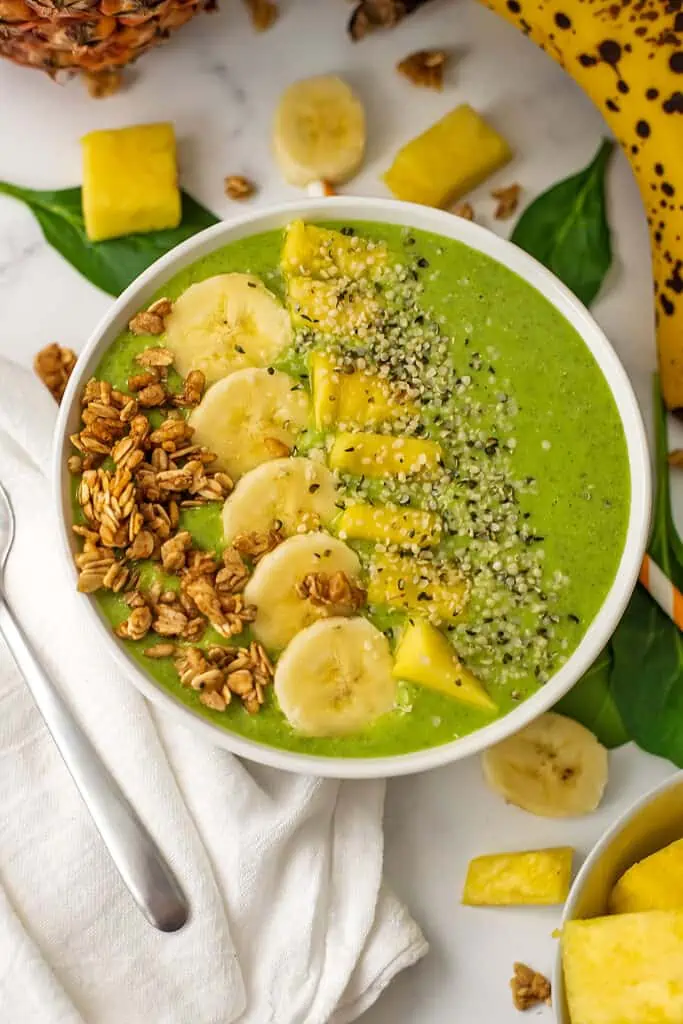 Pineapple banana spinach smoothie bowl with banana slices, pineapple chunks, granola and hemp hearts on top.