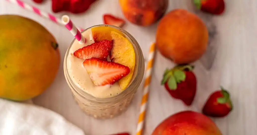 Strawberry peach mango smoothie surrounded by mangos, strawberries and peaches on a white table.