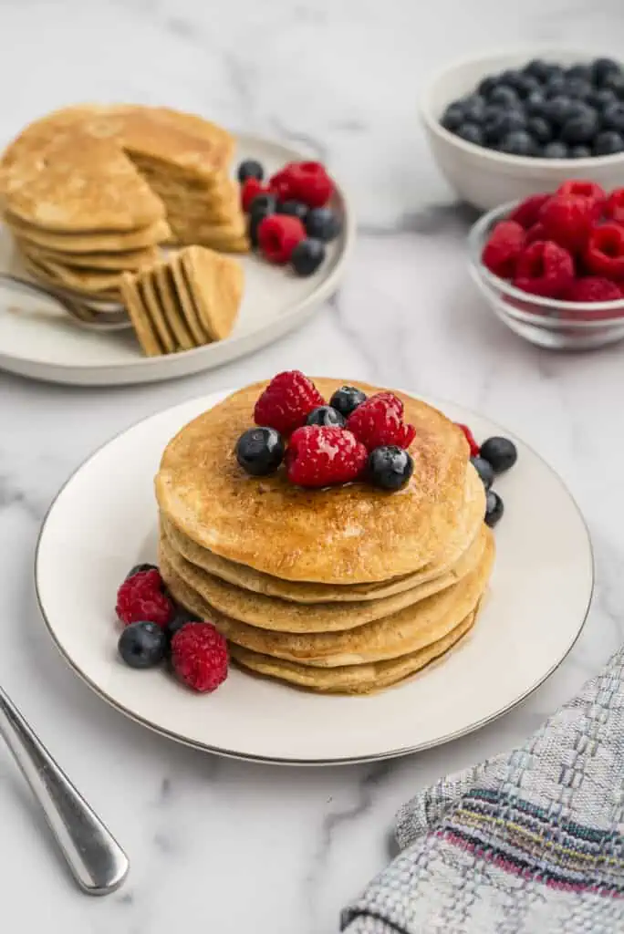 Gluten free oat flour pancakes with fruit on top with a stack of pancakes in the background.
