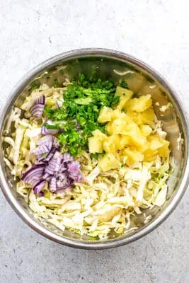 Pineapple, cilantro and red onion added to slaw in stainless steel bowl.
