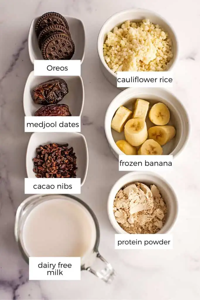 Ingredients to make oreo protein shake in ramekins on marble countertop.