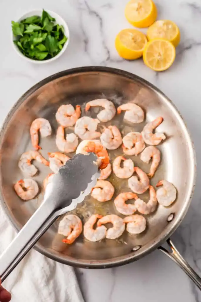 Tongs flipping shrimp in a stainless steel skillet.