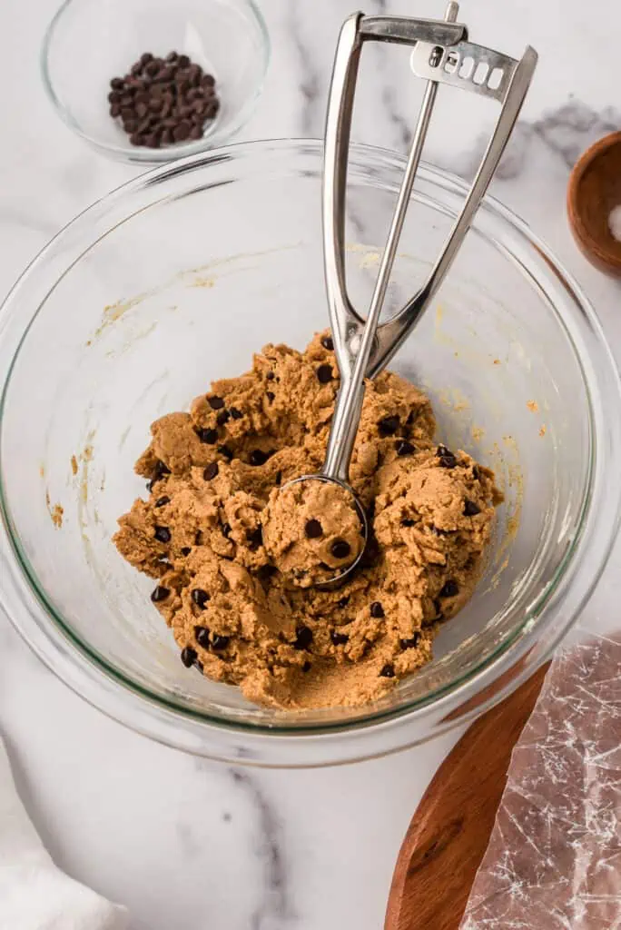 Protein almond butter ball mixture in a glass bowl with a cookie scoop resting in the bowl.