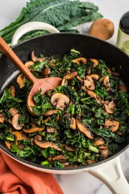 Wood spoon resting in skillet with sauteed kale and mushrooms.