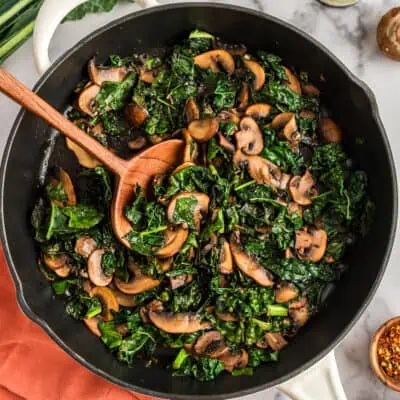 Wood spoon in a large skillet filled with balsamic kale and mushrooms.
