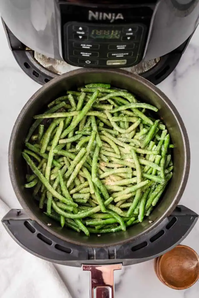 Frozen green beans with salt and garlic powder in air fryer basket. 
